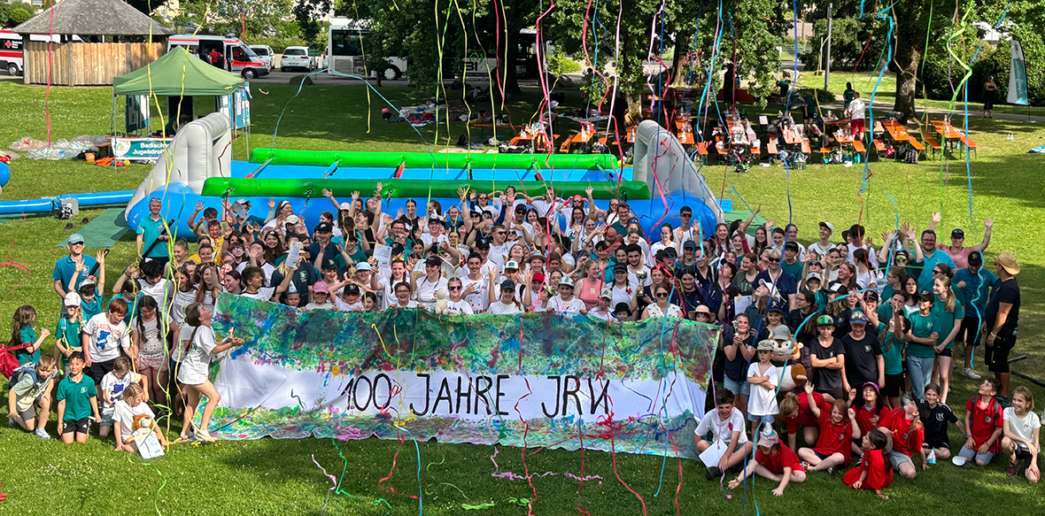 Eine große Menschengruppe versammelt sich auf einer Wiese und hält ein buntes Banner mit der Aufschrift "100 Jahre JRK". Vom Himmel fällt Konfetti runter.