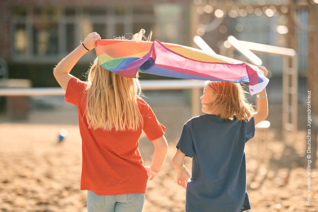 Zwei Kinder (eines mit rotem und eines mit petrolfarbenem T-Shirt) halten gemeinsam eine Regenbogen-Fahne über ihre Köpfe. Sie sind von hinten auf einem Spielplatz zu sehen.