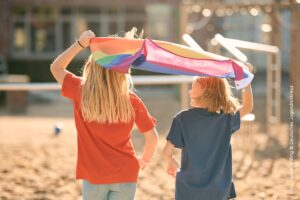 Zwei Kinder (eines mit rotem und eines mit petrolfarbenem T-Shirt) halten gemeinsam eine Regenbogen-Fahne über ihre Köpfe. Sie sind von hinten auf einem Spielplatz zu sehen.